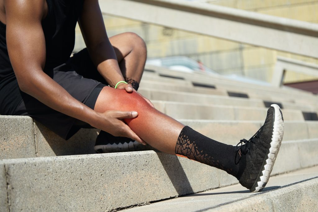 Junger Sportler mit kräftigen, athletischen Beinen, der sich das Knie hält, nachdem er beim Lauftraining eine Bänderverletzung erlitten hat. Er sitzt auf einer Treppe im Freien und reibt sich die wunde Stelle mit einem schmerzhaften Gesichtsausdruck ein.